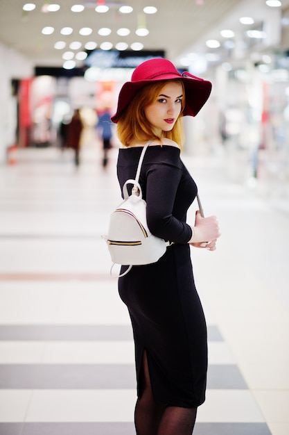 Menina de cabelos vermelha moda vestir vestido preto e chapéu vermelho com mochila senhoras posou no comércio shopping center.