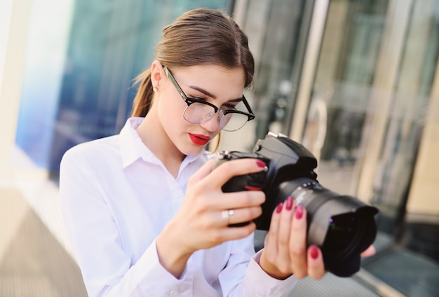 Menina de camisa branca, segurando nas mãos a câmera pressiona o botão do obturador e olha para o visor. Fotografia, fotógrafo, paparazzi