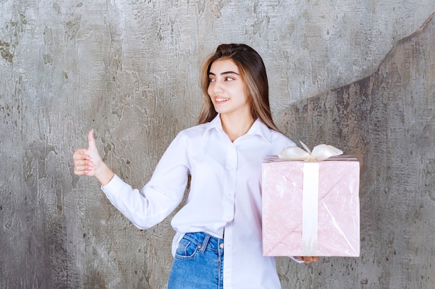 Menina de camisa branca segurando uma caixa de presente rosa embrulhada com fita branca e mostrando sinal positivo