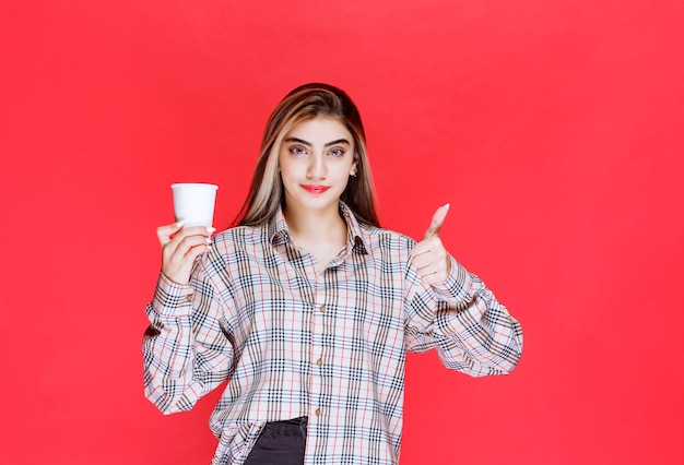 Menina de camisa xadrez segurando uma xícara de café descartável branca e apreciando o sabor