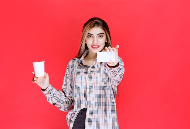 Menina de camisa xadrez segurando uma xícara de café e apresentando seu cartão de visita