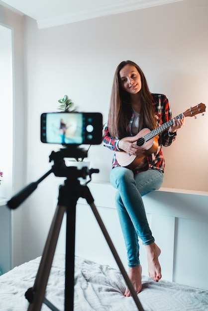 Menina de camiseta e calça jeans escreve um vlog no telefone sobre tocar ukulele.