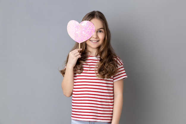 Menina de camiseta listrada cobrindo o olho com coração rosa na vara mostrando sentimentos românticos