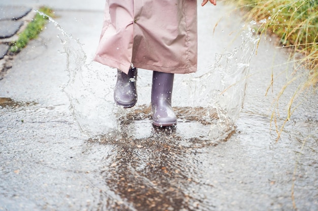 Menina de capa de chuva rosa impermeável botas de borracha roxa saltos engraçados através de poças na estrada de rua em clima de dia chuvoso Primavera outono Diversão infantil depois da chuva Atividade de recreação ao ar livre