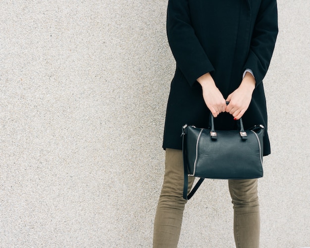 Menina de casaco preto, jeans verde e uma bolsa na mão fica em uma superfície de parede bege