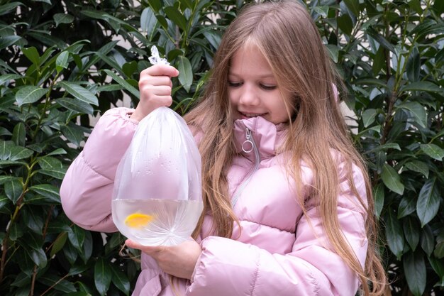 Menina de casaco rosa sobre fundo de folhagem verde segurando pequenos peixes dourados em saco plástico