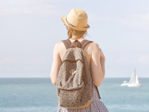 Menina de chapéu com uma mochila em pé no litoral. Veleiro à distância. Vista traseira