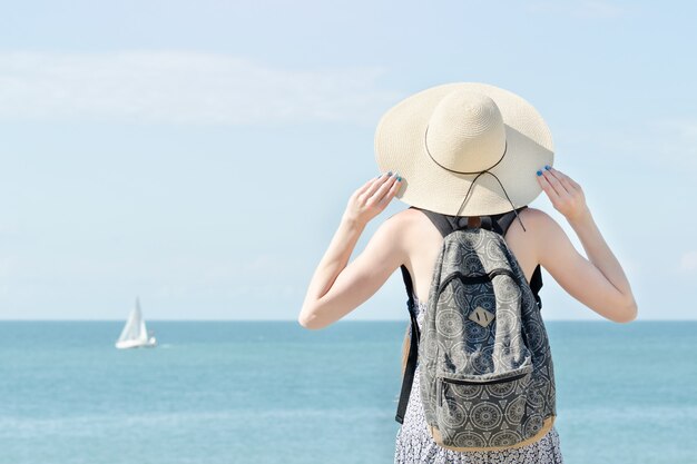 Menina de chapéu com uma mochila em pé no litoral.