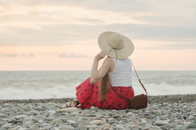 Menina de chapéu senta-se em uma praia de calhau.