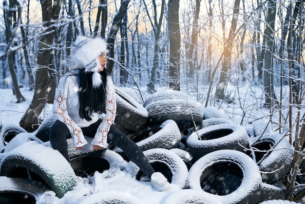 Menina de chapéu sentado em pneus de carro na floresta no inverno.