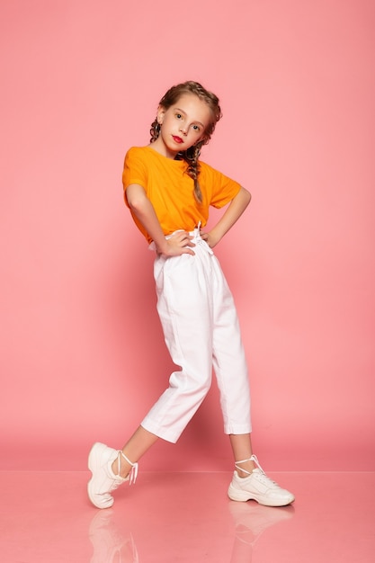 Menina de comprimento total na parede rosa do estúdio. vestindo uma camiseta laranja e calça branca e tênis branco.