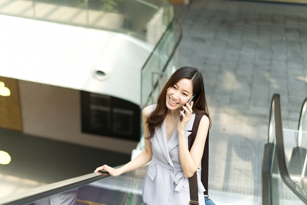 Menina de escritório asiática que fala no telefone móvel que está no elevador no shopping.