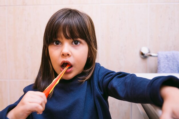 Menina de moletom azul escuro, escovando os dentes, no banheiro. Conceito de escovação, higiene e infância.