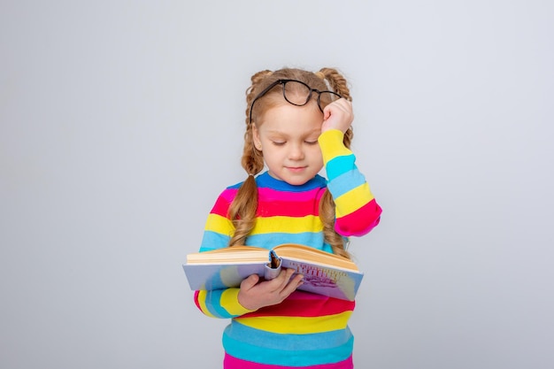 menina de óculos de suéter multicolorido está segurando um livro sobre um fundo branco fazendo caretas