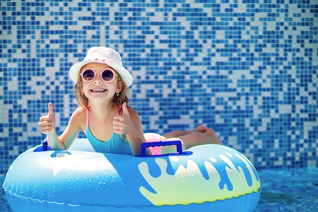 Menina de óculos escuros e chapéu com unicórnio no anel amarelo inflável na piscina do resort de luxo em férias de verão na ilha de praia tropical.