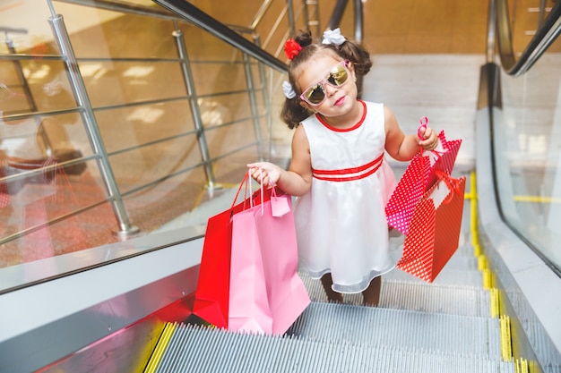 Menina de óculos escuros em uma escada rolante em um shopping com bolsas coloridas