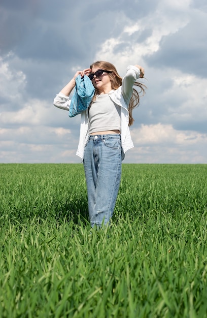 menina de óculos escuros endireita o cabelo em um campo de grama verde, no contexto de um céu nublado.