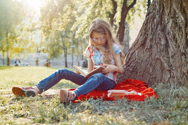 Menina de óculos lendo livro no parque, na grama perto da árvore