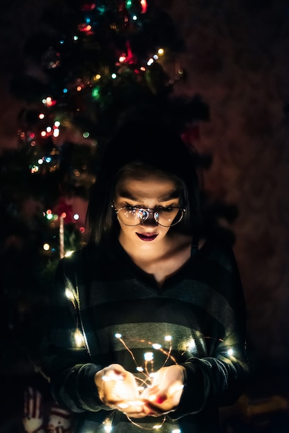 Foto menina de óculos, segurando uma guirlanda com luzes à noite em casa