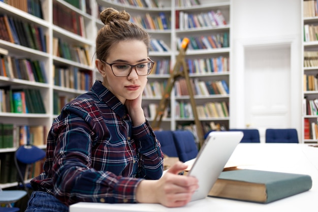 Menina de óculos, trabalhando com tablet sentado na biblioteca.