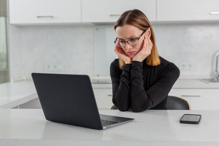 Menina de óculos tristemente olha para o monitor do laptop sentada em casa na cozinha branca foto de alta qualidade