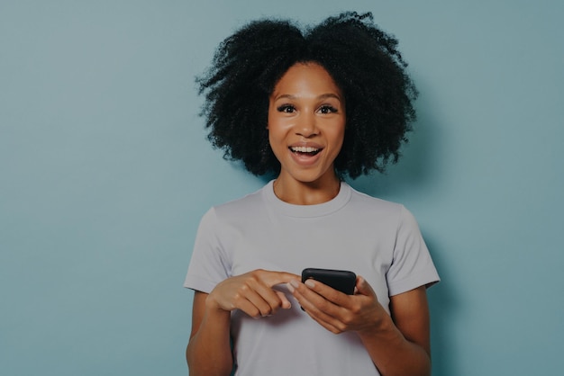 Menina de pele escura feliz segurando smartphone moderno e conversando com um amigo online ou usando aplicativo móvel apontando para a tela do dispositivo e olhando para a câmera com expressão de rosto animado