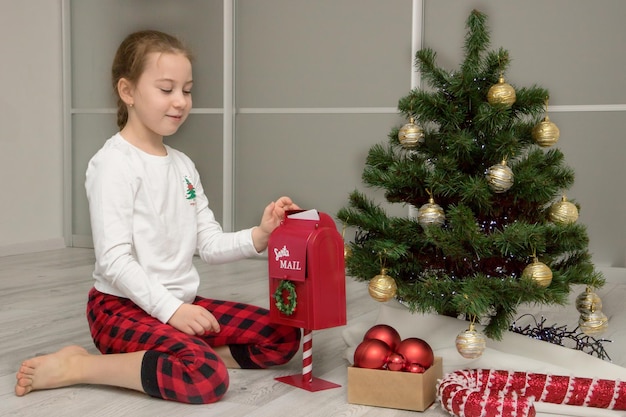 Menina de pijama perto da árvore de Natal joga uma carta para o Papai Noel na caixa de correio presentes de Natal