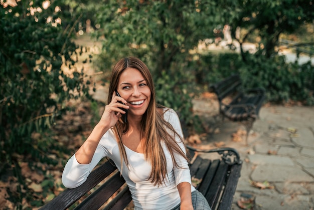 Menina de sorriso atrativa que senta-se em um banco no parque e que fala no telefone esperto.