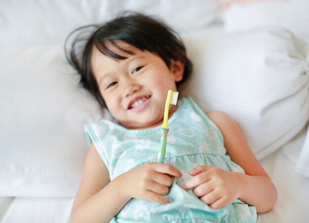 Menina de sorriso da criança que escova os dentes na cama na manhã, foco em sua cara.