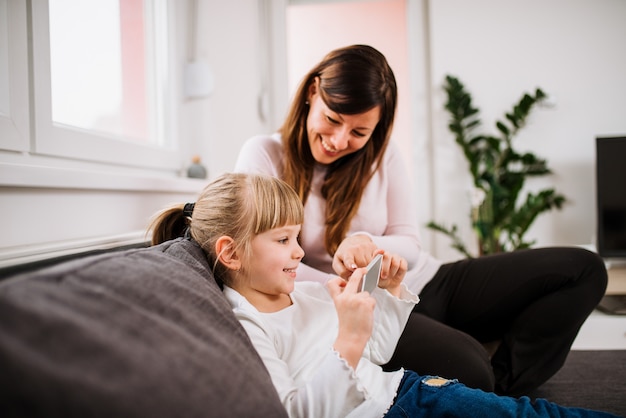 Menina de sorriso e sua mãe que usa o smartphone.