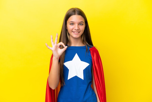 Menina de super-herói caucasiana isolada em fundo amarelo, mostrando sinal de ok com os dedos