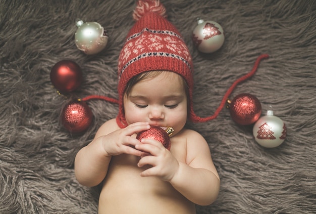 Menina de um ano deitada em uma colcha, de chapéu vermelho, brincando com os brinquedos de ano novo
