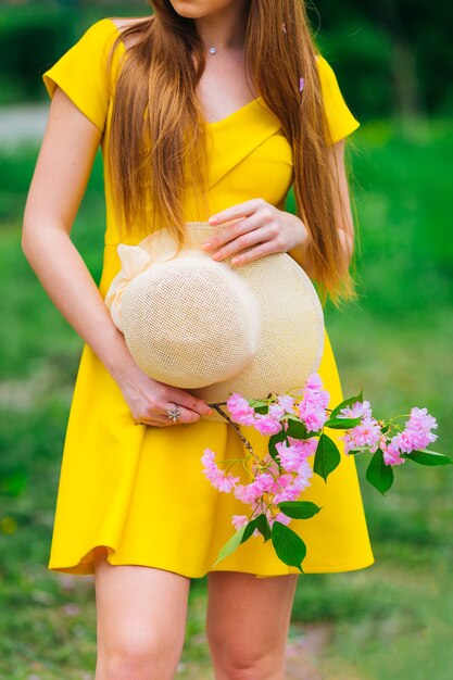 Menina de vestido amarelo com cabelo comprido segurando um chapéu e um galho com flores cor de rosa