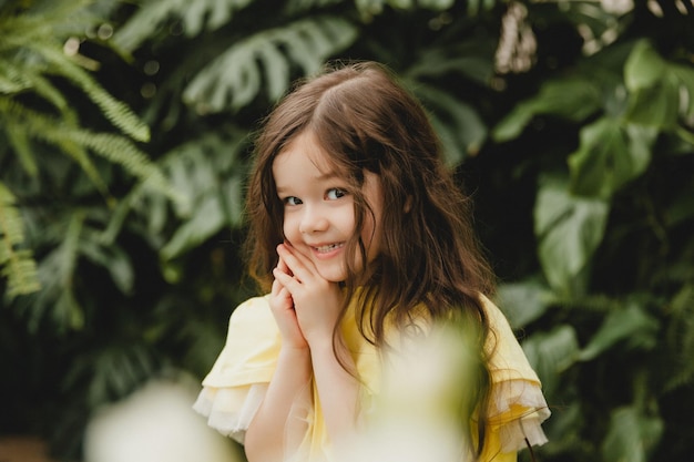 Menina de vestido amarelo em um jardim botânico, uma criança fica perto das folhas de Monstera