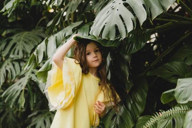Menina de vestido amarelo em um jardim botânico, uma criança fica perto das folhas de Monstera