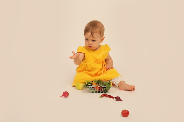 menina de vestido amarelo está sentada sobre um fundo branco ao lado de uma pequena cesta com legumes