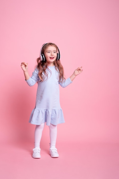 Menina de vestido azul ouvindo música dançando em grandes fones de ouvido brancos no fundo rosa