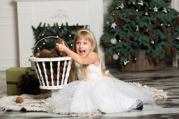 Menina de vestido branco brinca com pinhas em decorações de Natal, árvore de Natal e lareira.