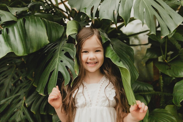 Menina de vestido branco em um jardim botânico, uma criança fica perto das folhas de Monstera