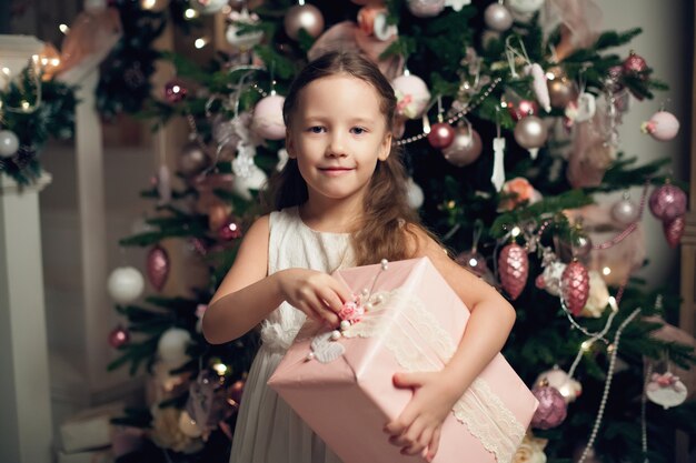 Menina de vestido em pé perto da árvore de Natal e segurando o presente