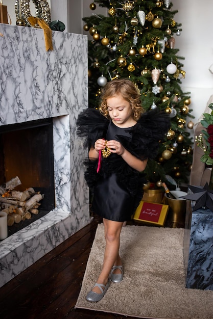 Menina de vestido preto com presente de Natal nas mãos perto de árvores de Natal com luzes Feliz Natal e Boas Festas