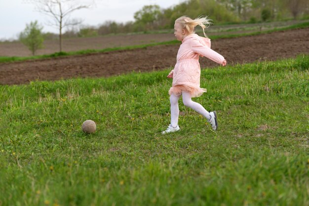 Menina de vestido rosa corre pelo prado verde e chuta bola Criança brinca no campo na primavera