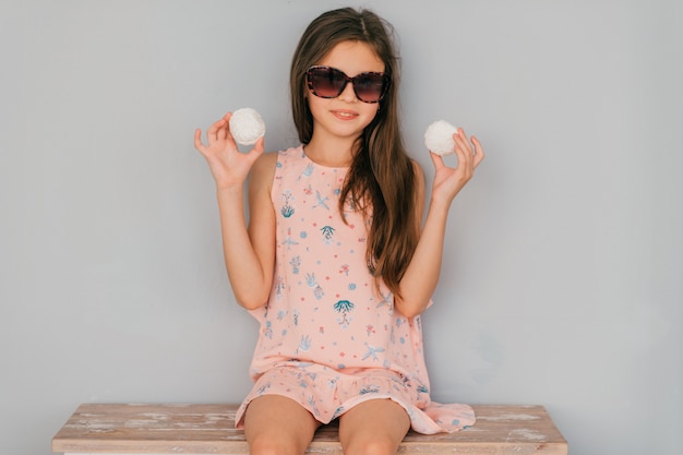 Menina de vestido rosa, sentado em óculos de sol com marshmallows nas mãos contra a parede cinza