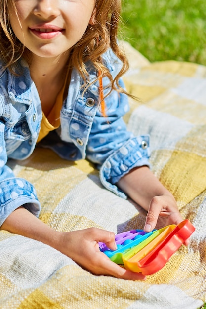 Menina deita-se em um cobertor na grama ao ar livre e joga pop, mãos de criança jogando bolhas de um brinquedo de destruição de arco-íris, brinquedo de agitação no quintal da casa em um dia ensolarado de verão, férias de verão.