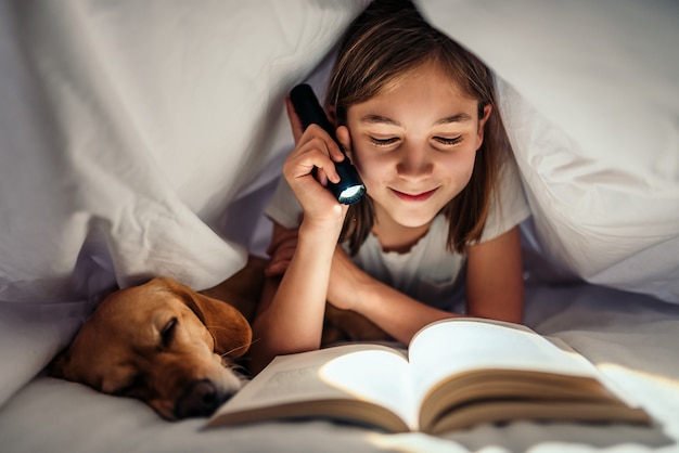 Menina deitada na cama com seu cachorro sob o livro de leitura geral tarde da noite