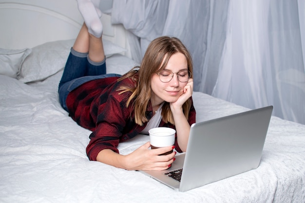 Menina deitada na cama com um laptop e tomando um café