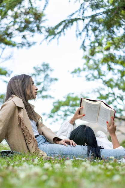Menina deitada nas pernas da amiga enquanto lê um livro na grama Estilo de vida de uma menina deitada nas pernas da amiga lendo um livro no campo
