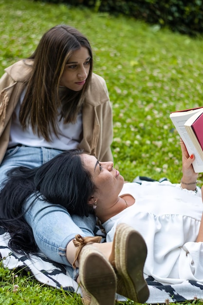 Menina deitada nas pernas da amiga enquanto lê um livro na grama Estilo de vida de uma menina deitada nas pernas da amiga lendo um livro no campo