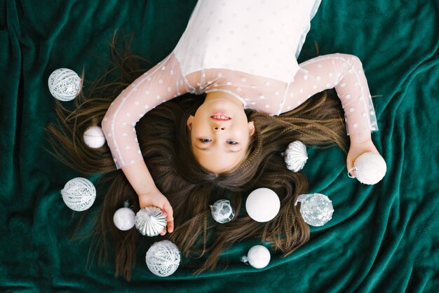 Menina deitada no chão em um cobertor, bolas brancas de Natal, ao lado da árvore de Natal, vista de cima, conceito de ano novo