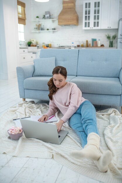 Menina deitada sobre uma manta no chão, encostada na mão, olhando para um laptop, ao lado de um prato com marshmallows.